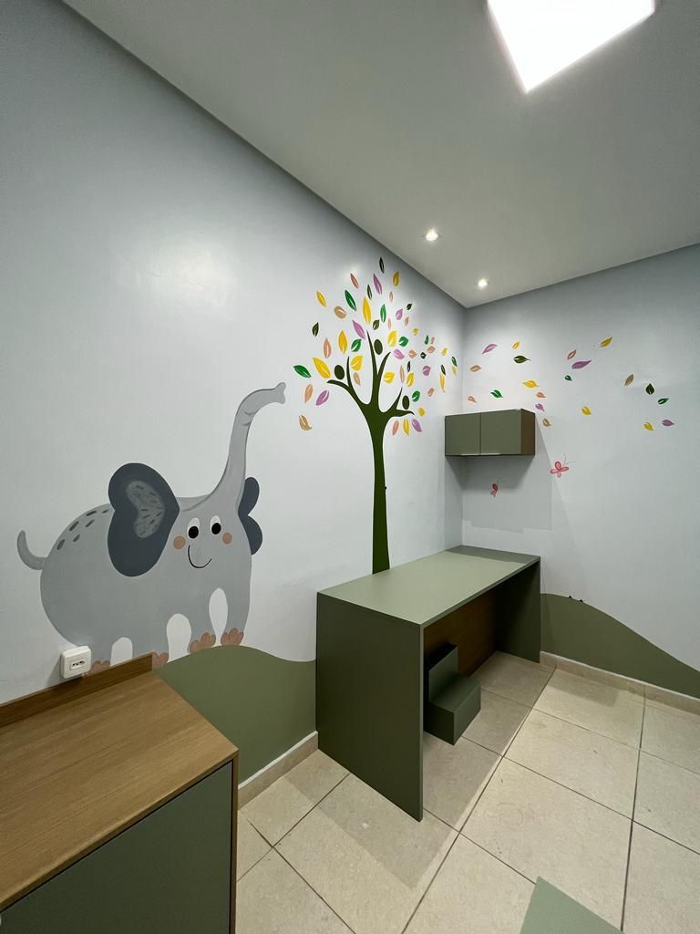 Consultório pediátrico com bancada verde, armários e mural de um elefante ao lado de uma árvore de folhas coloridas